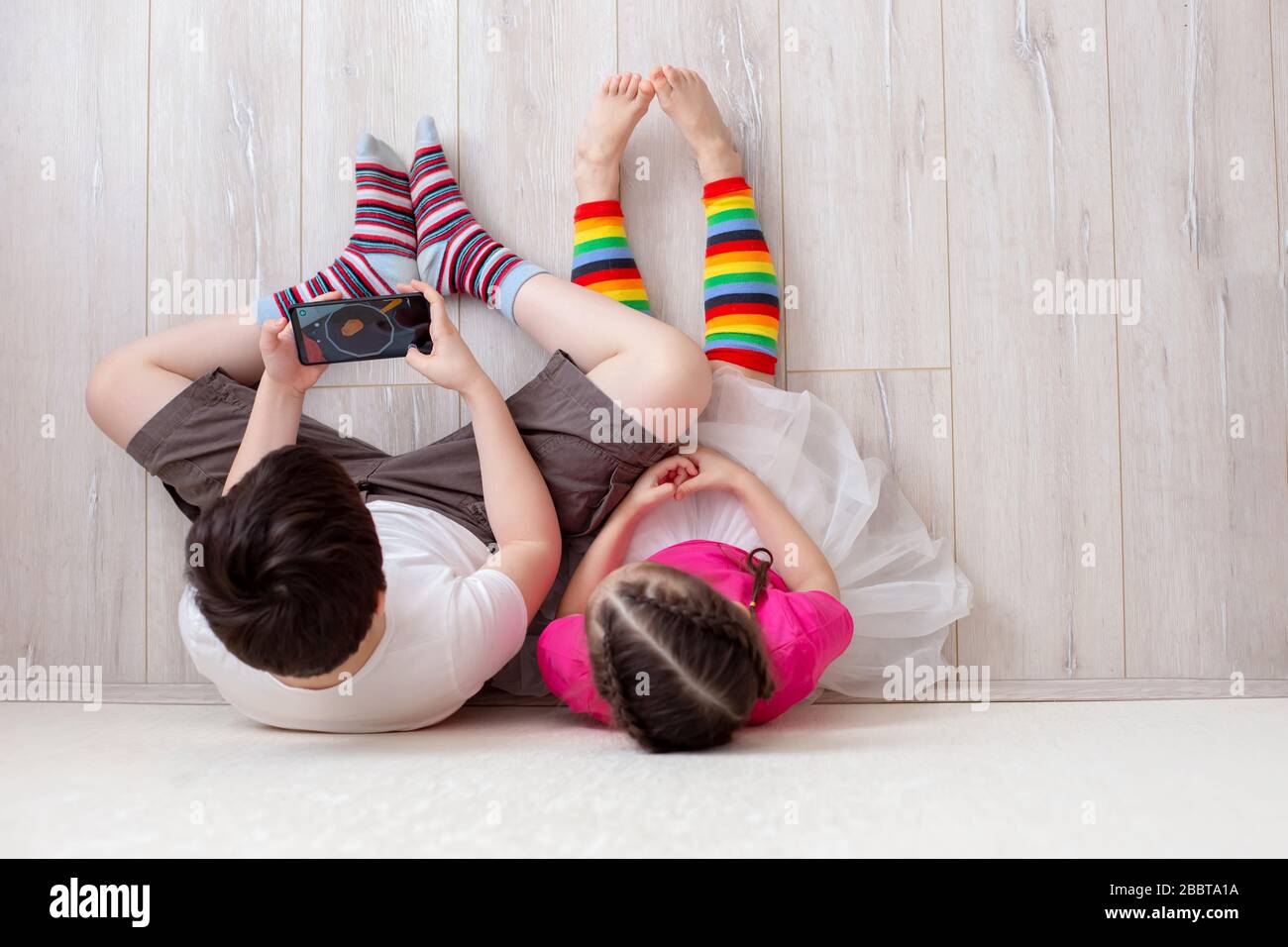 Dos niños, hermano y hermana, están sentados en el suelo contra la pared,  las piernas con calcetines largos a rayas brillantes y jugando con un  smartphone. Vista superior Fotografía de stock -