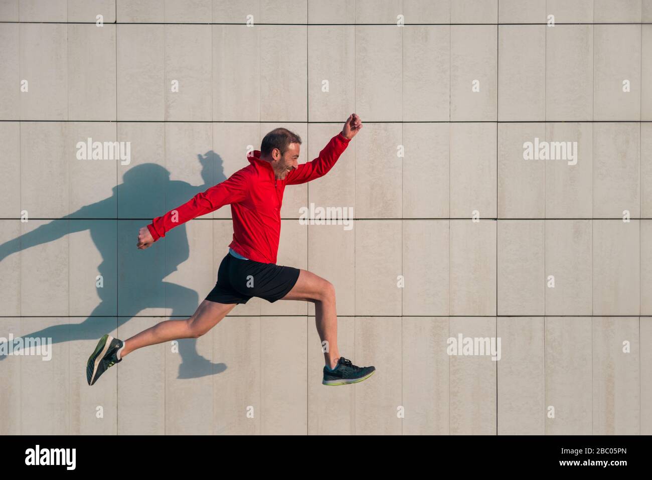 Atleta corriendo y saltando en un día soleado con su sombra en una pared blanca. Foto de stock