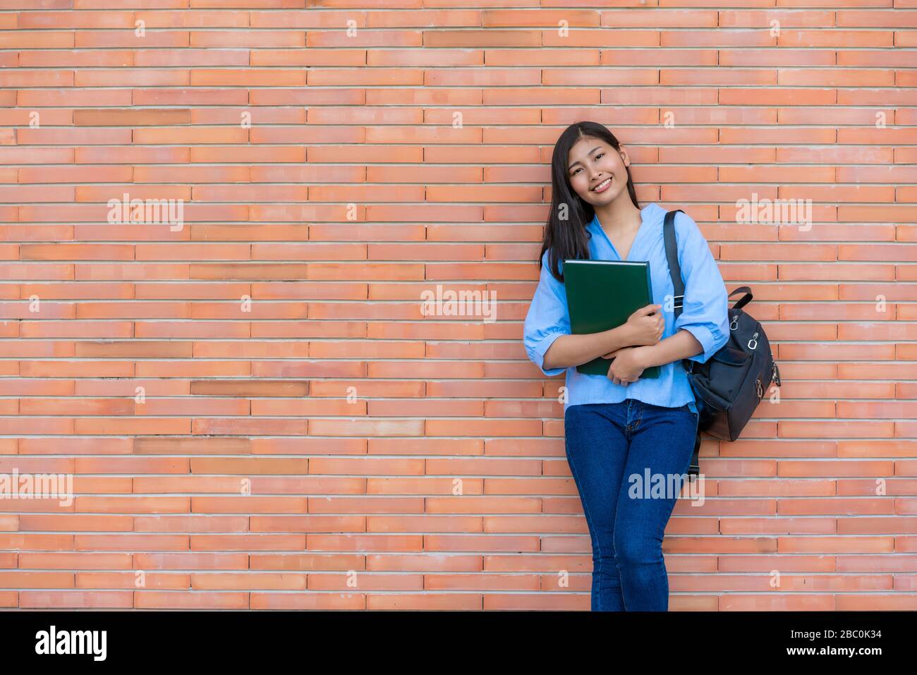 Una mujer sonriente asiática que sostiene un libro posando sobre fondo de ladrillo en el campus. Feliz niña estudiante de secundaria al aire libre. Educación, Aprendizaje, Stud Foto de stock