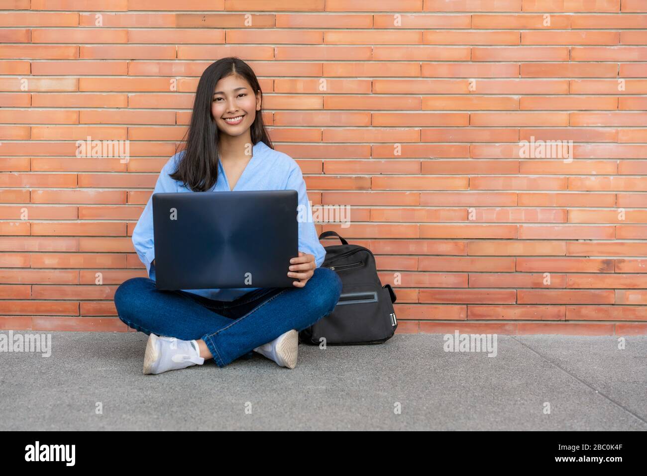 Mujer sonriente asiática estudiante sentado y usando el ordenador portátil sobre fondo de ladrillo en el campus. Feliz niña estudiante de secundaria al aire libre. Educación, aprendizaje, Foto de stock