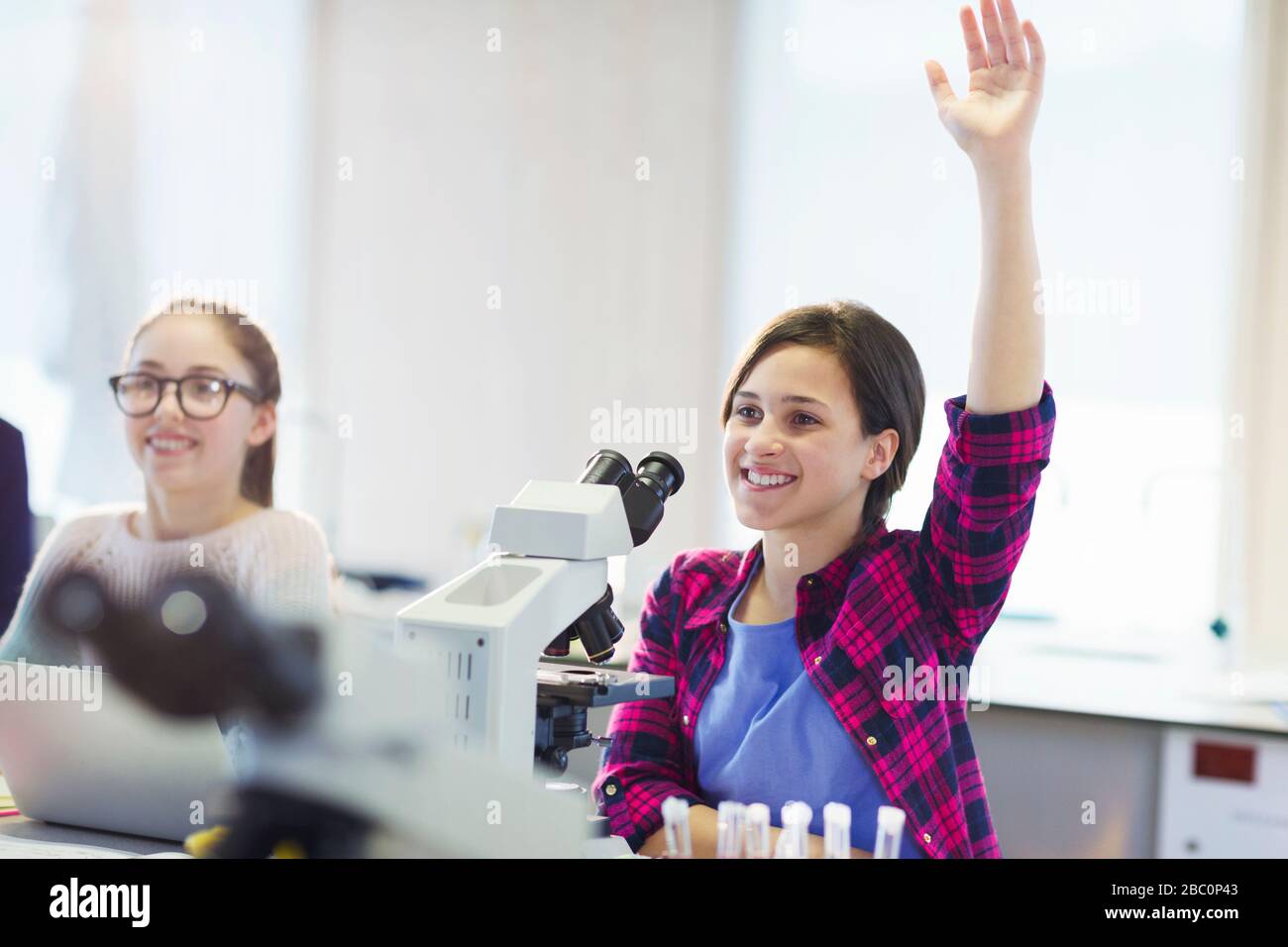 Estudiante sonriente haciendo una pregunta detrás del microscopio en el laboratorio del salón de clases Foto de stock