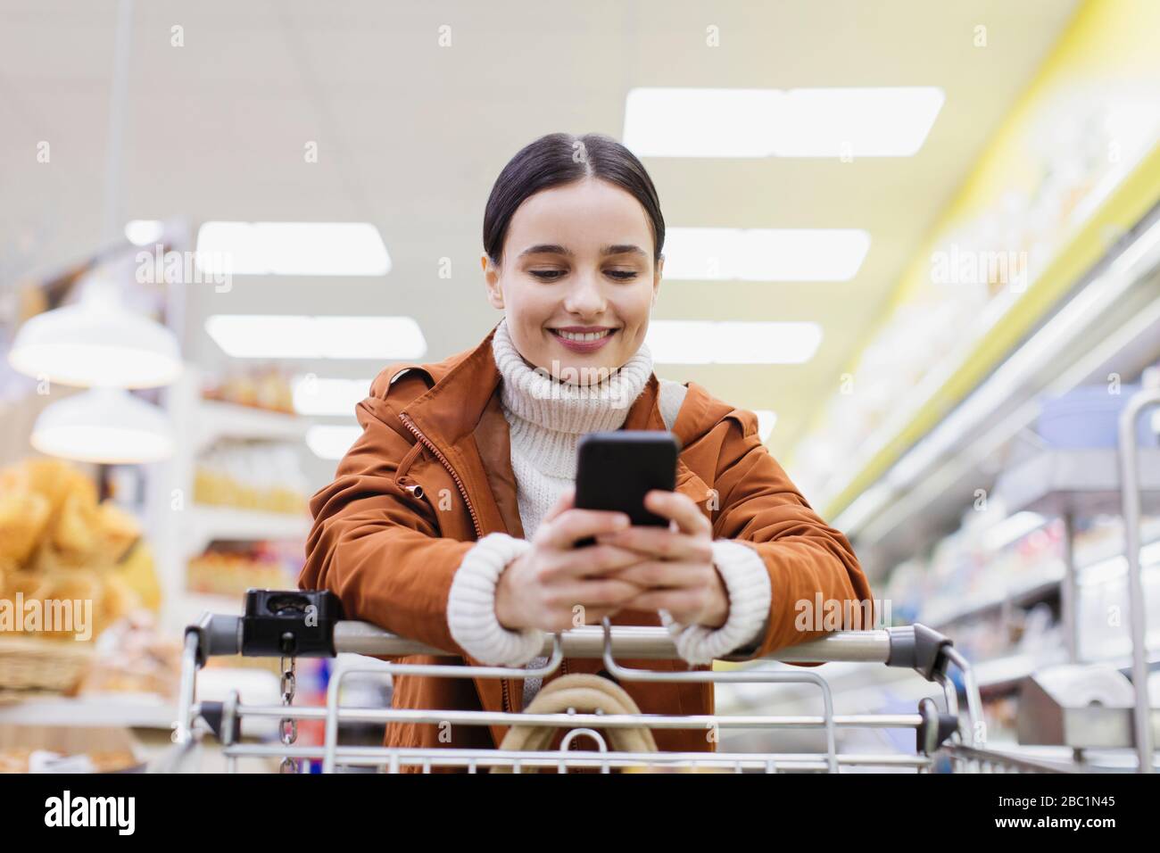 Mujer con el teléfono inteligente empujando carro de compras en el supermercado Foto de stock
