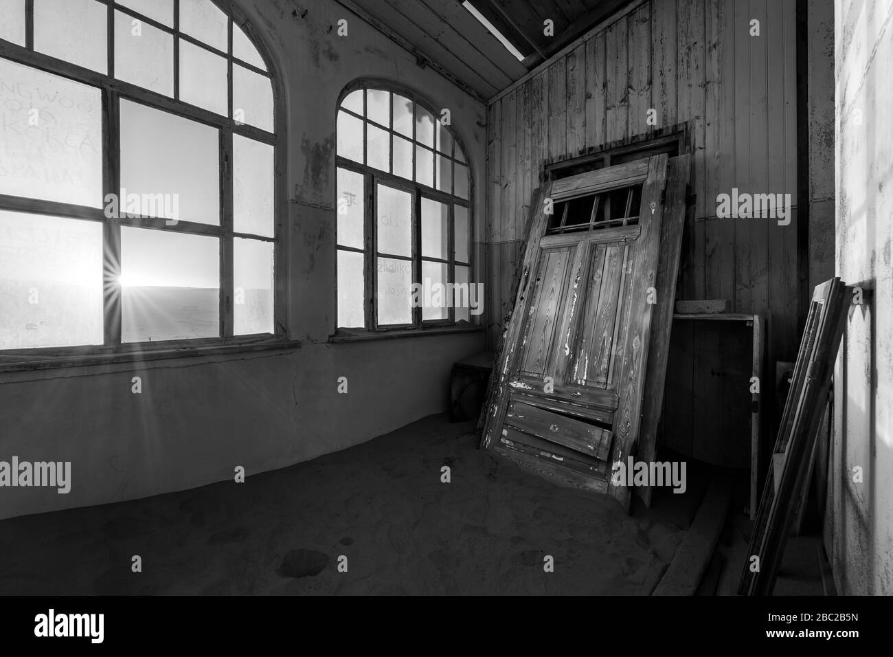 Una dramática fotografía en blanco y negro dentro de una casa abandonada al amanecer, con un estallido de sol a través de ventanas rotas y una puerta enterrada en la arena del desierto, t Foto de stock