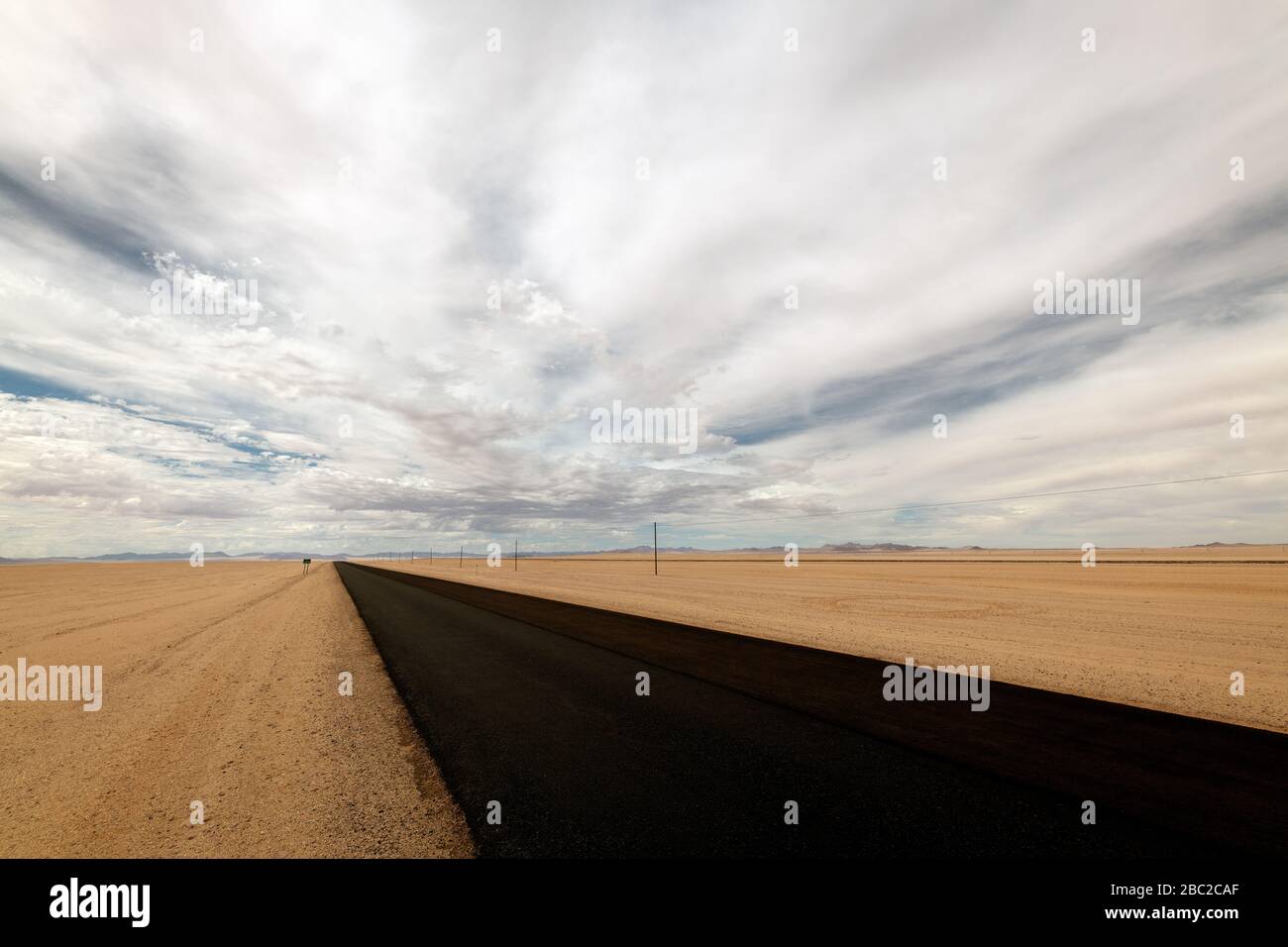 Un paisaje desértico espeluznante tomado cerca de Luderitz, Namibia, con la carretera nacional de alquitrán negro que conduce a la distancia, la arena del desierto en el primer plano y Foto de stock