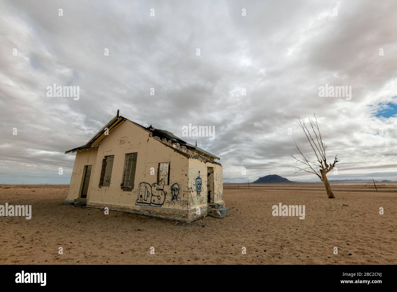 Un espeluznante paisaje desértico cerca de Luderitz, Namibia, con una antigua casa abandonada cubierta de grafitti y un árbol muerto contra un oscuro, tormentoso y el mo Foto de stock