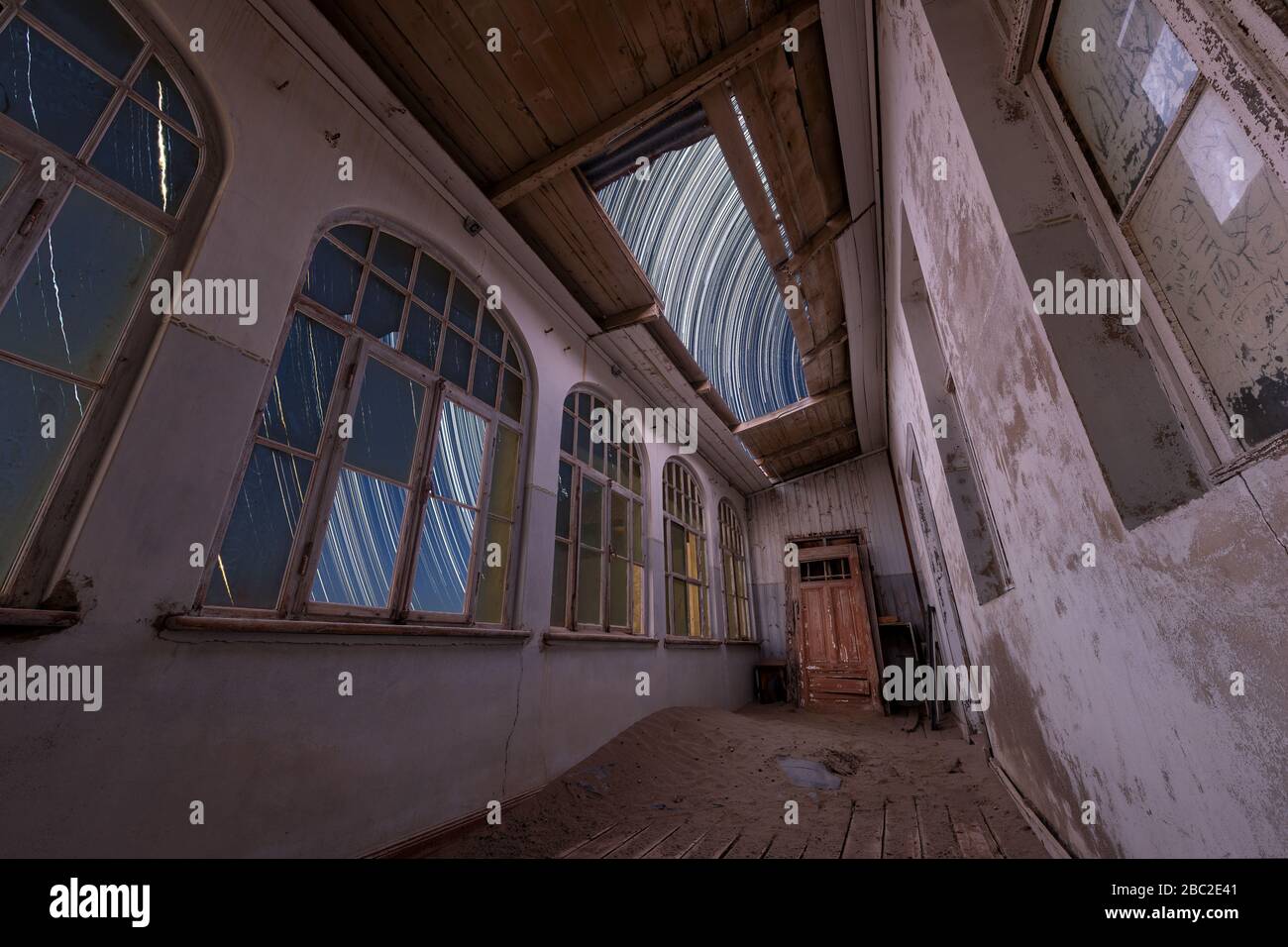 Un espectacular cielo nocturno en el interior de un edificio abandonado con senderos estelares que aparecen a través del techo roto y la arena del desierto y una puerta dentro de la habitación, Foto de stock