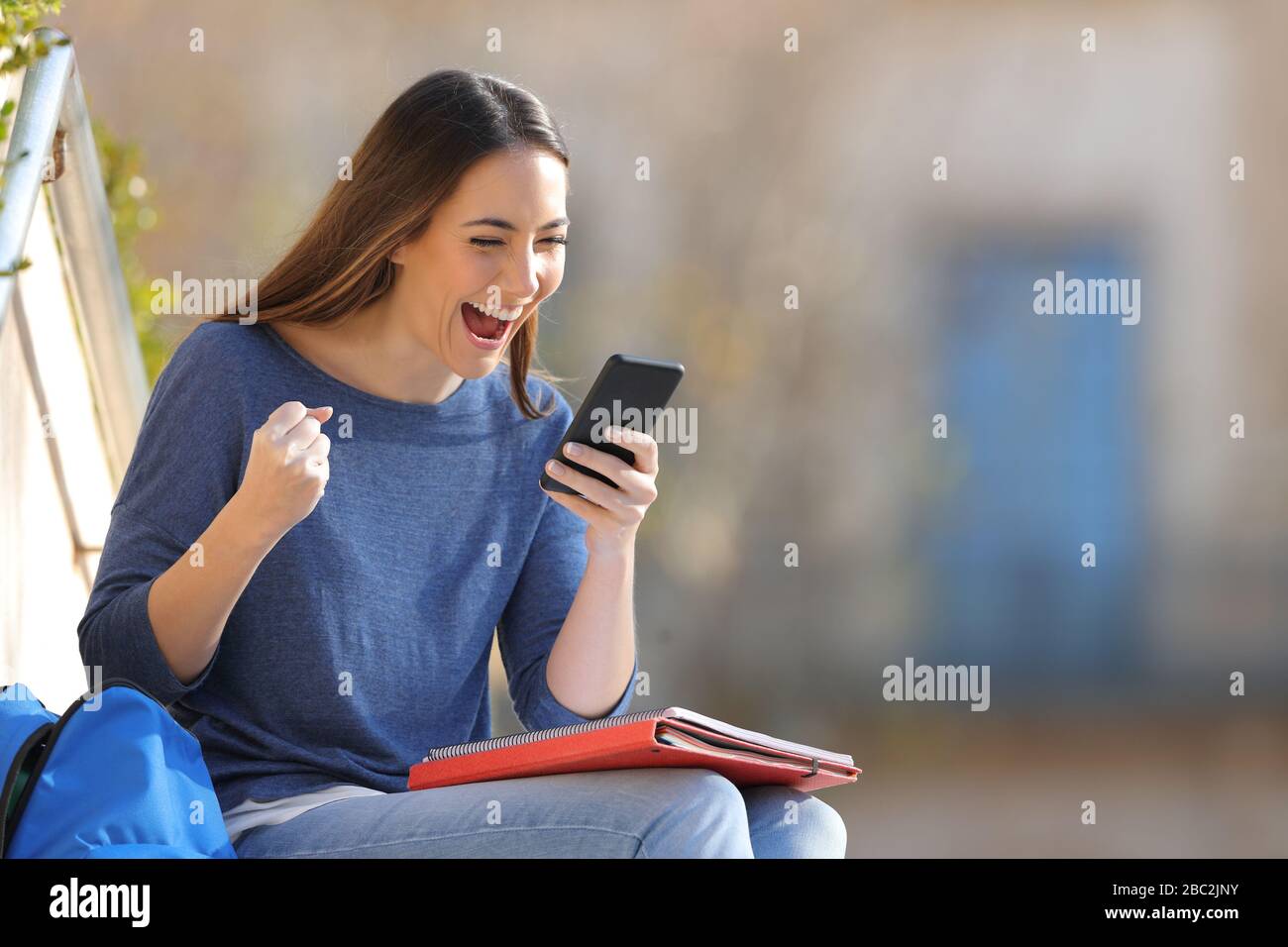 Un estudiante emocionado comprobando el contenido del teléfono inteligente sentado al aire libre en un campus universitario Foto de stock