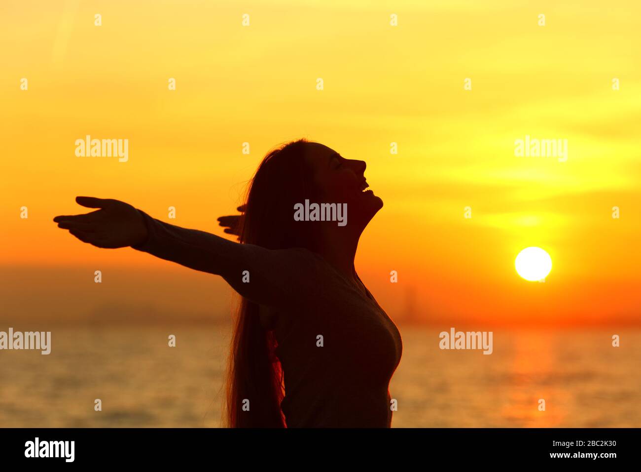 Vista lateral retrato de una mujer feliz celebrando un nuevo día estirando los brazos al amanecer en la playa Foto de stock
