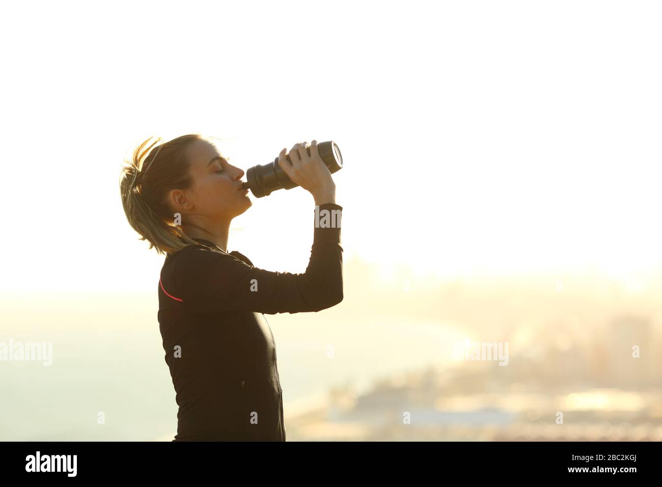 Vista lateral retrato de una mujer corredora que bebe agua de una botella hidratada después de hacer ejercicio en las afueras de la ciudad Foto de stock