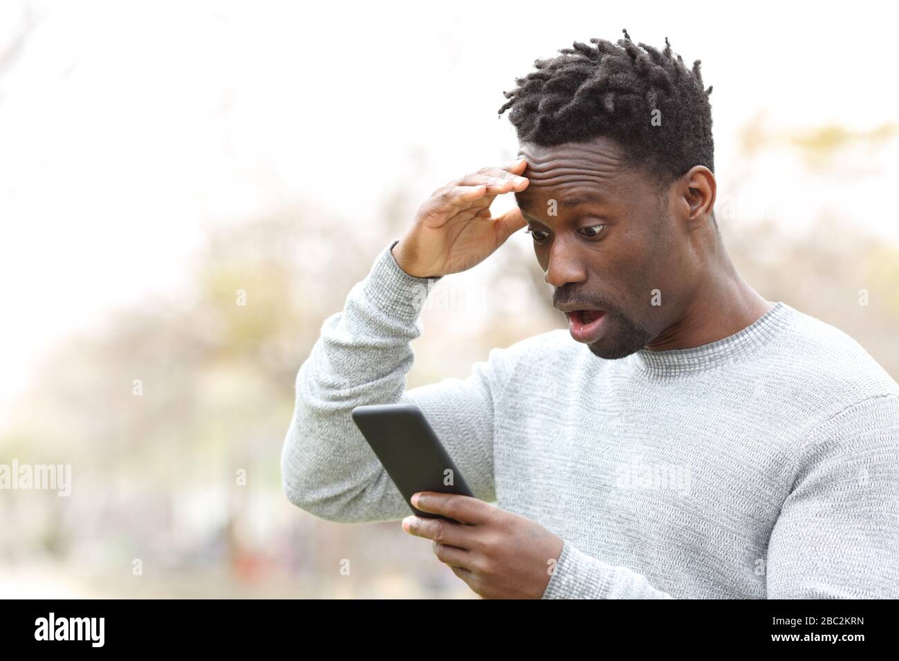 Sorprendido hombre negro comprobando teléfono móvil increíble noticias de pie en un parque Foto de stock