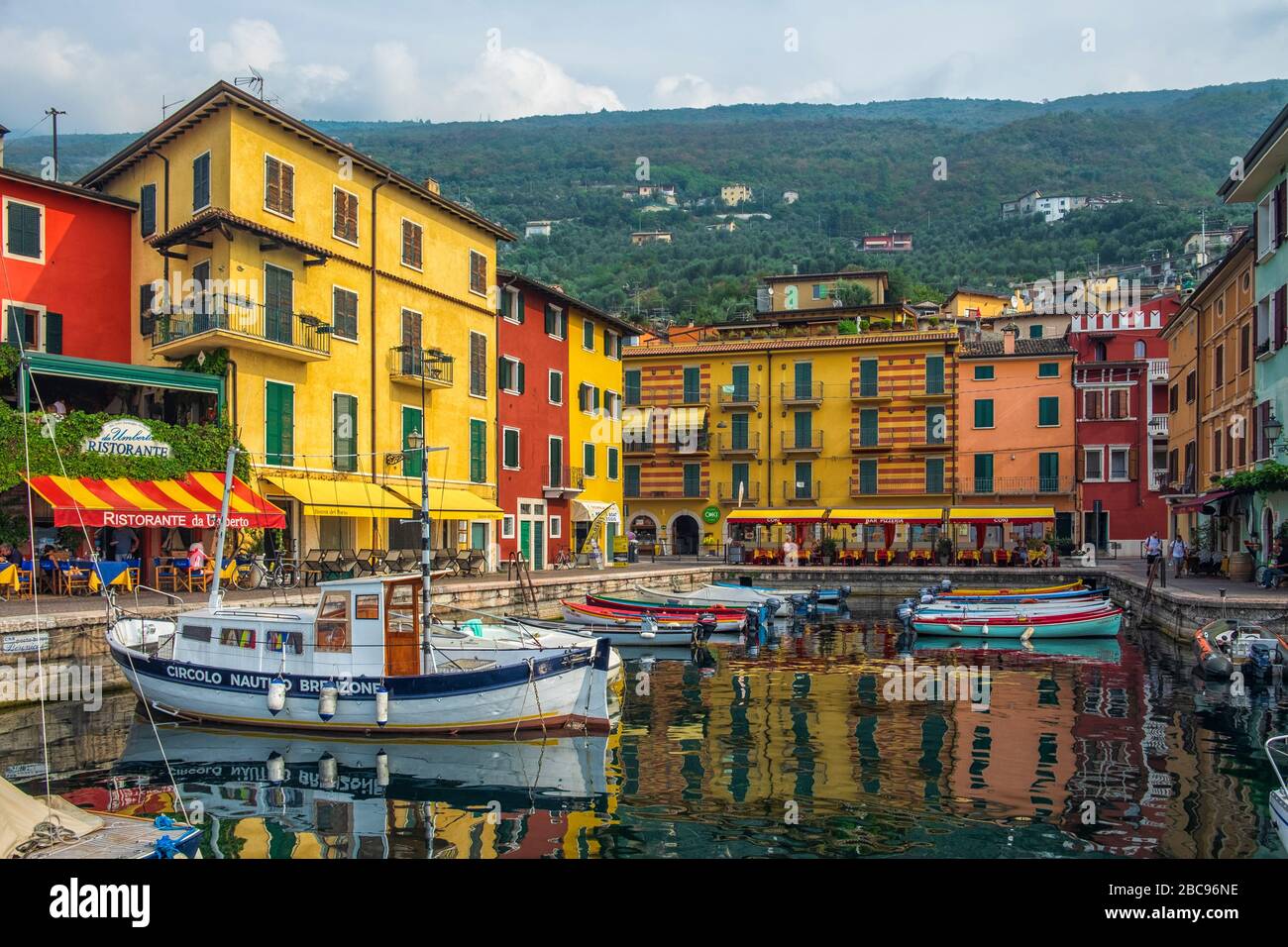 Paseo marítimo del lago Garda con casas, turistas y barcos en Torbole, Italia Foto de stock