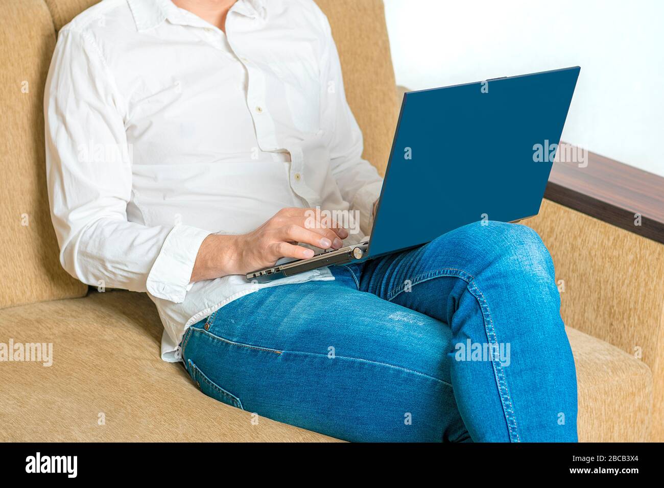 Hombre joven en camisa blanca jeans azules el portátil, escribe en el portátil, mirando en la pantalla blanca en la en casa sentado en un acogedor sofá beige