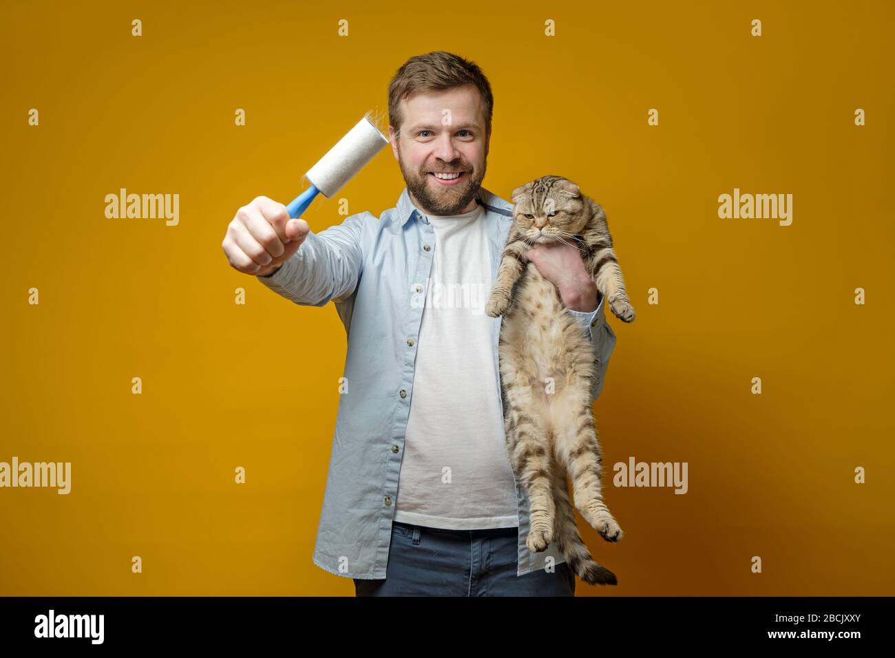 El hombre contento y sonriente sostiene en su mano un rodillo con cinta  adhesiva en el pelo de los animales, en el otro se jadera con su amado gato  Fotografía de stock -