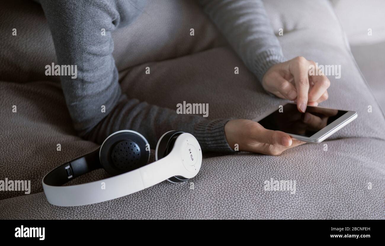 La mujer está acostada al teléfono en el sofá con auriculares a su lado. Permanezca en casa campaña de las redes sociales para la prevención del coronavirus. Foto de stock