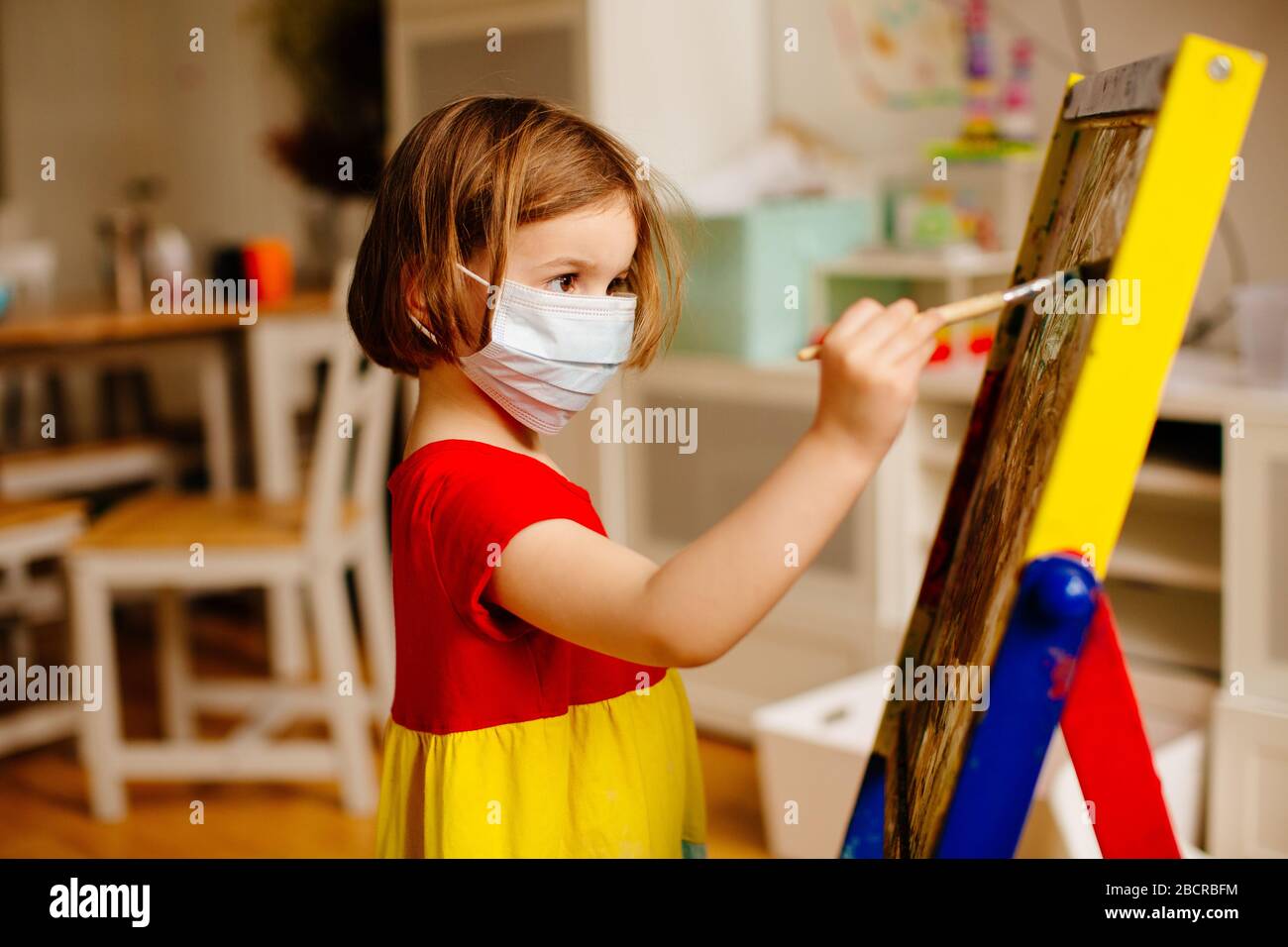 Retrato de un pequeño niño preescolar con máscara facial contra el coronavirus, pintando en una caballete de arte en casa Foto de stock