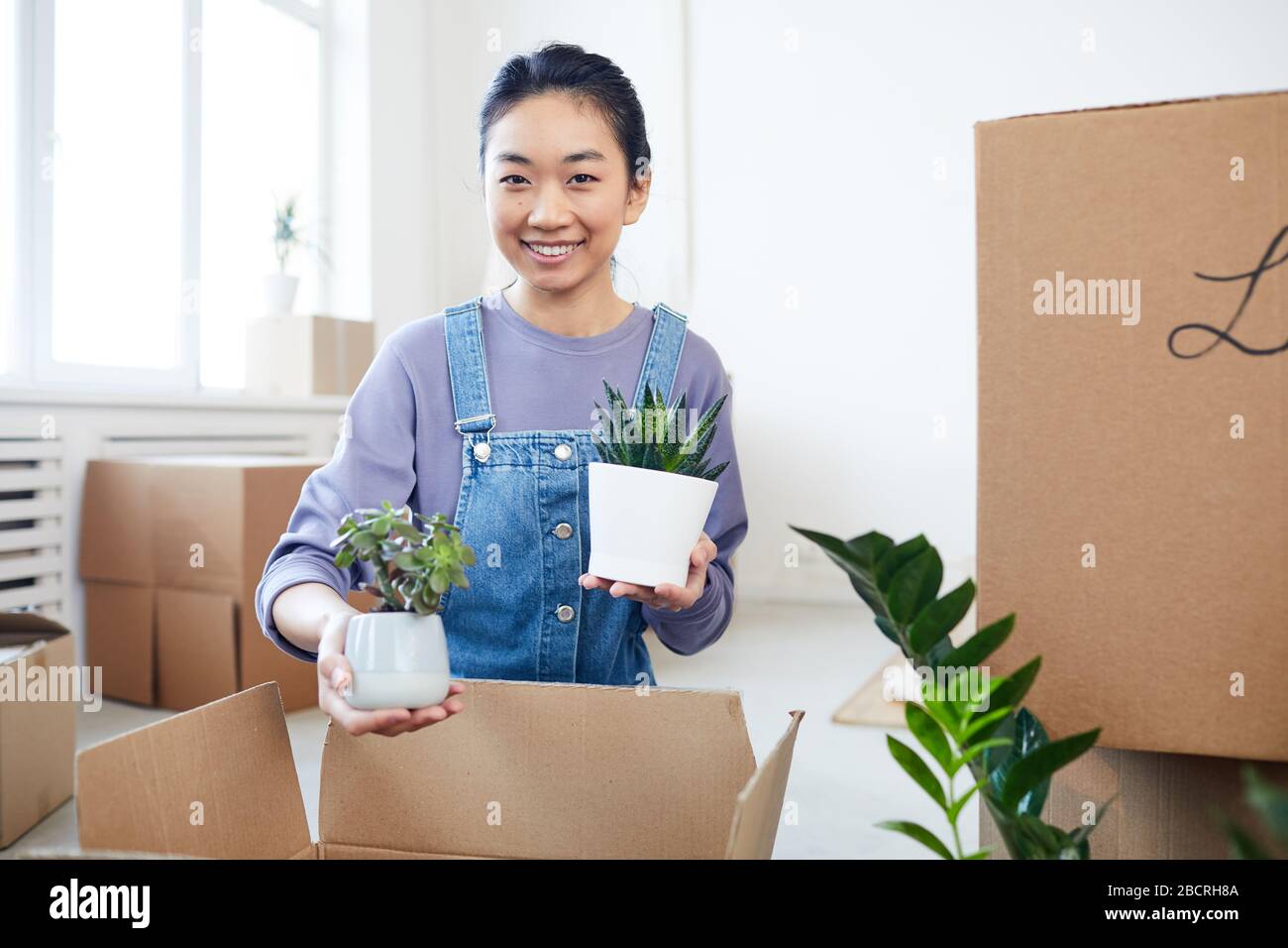 Retrato de una joven mujer asiática empaquetando plantas en cajas de cartón y sonriendo con la cámara mientras se mueve a una nueva casa o apartamento, copiar espacio Foto de stock