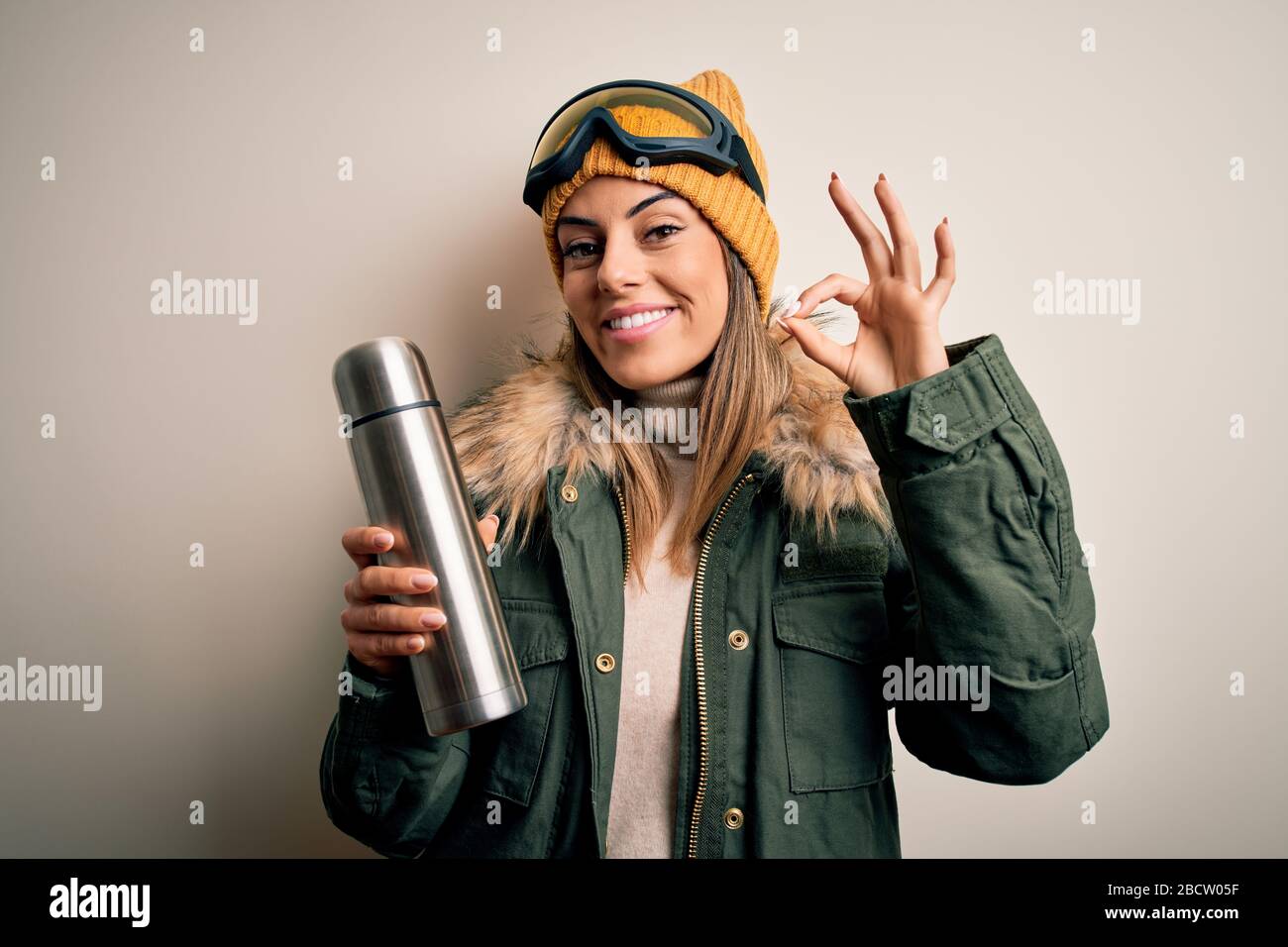 Joven hermosa morena skier mujer usando gafas de esquí tomando termo con café haciendo ok signo con los dedos, excelente símbolo Foto de stock