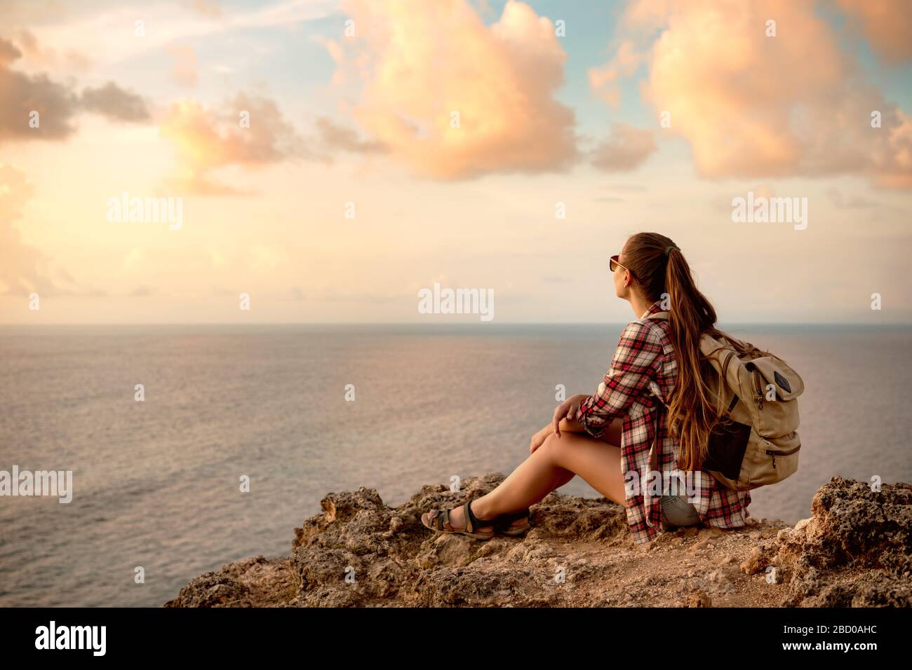 Una chica con mochila se sienta en el acantilado contra el mar y mira a la puesta de sol Foto de stock
