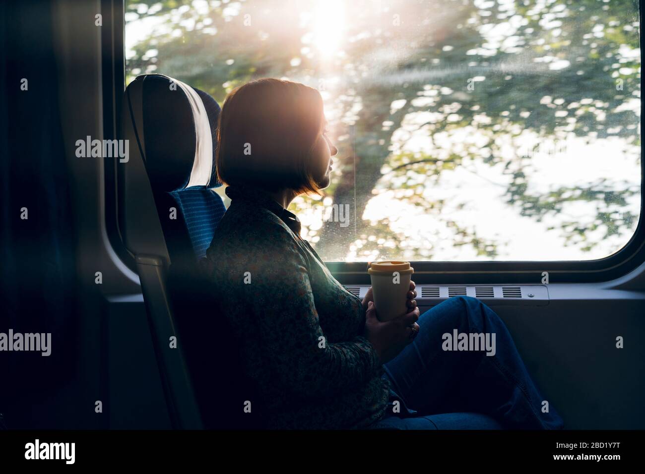 Una mujer sentada por la ventana de un tren de pasajeros con un portátil y una taza de café Foto de stock