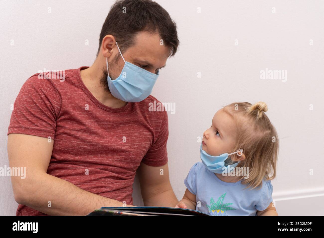 Niña en máscara médica mira al padre que está en máscara facial y leyendo el libro Foto de stock
