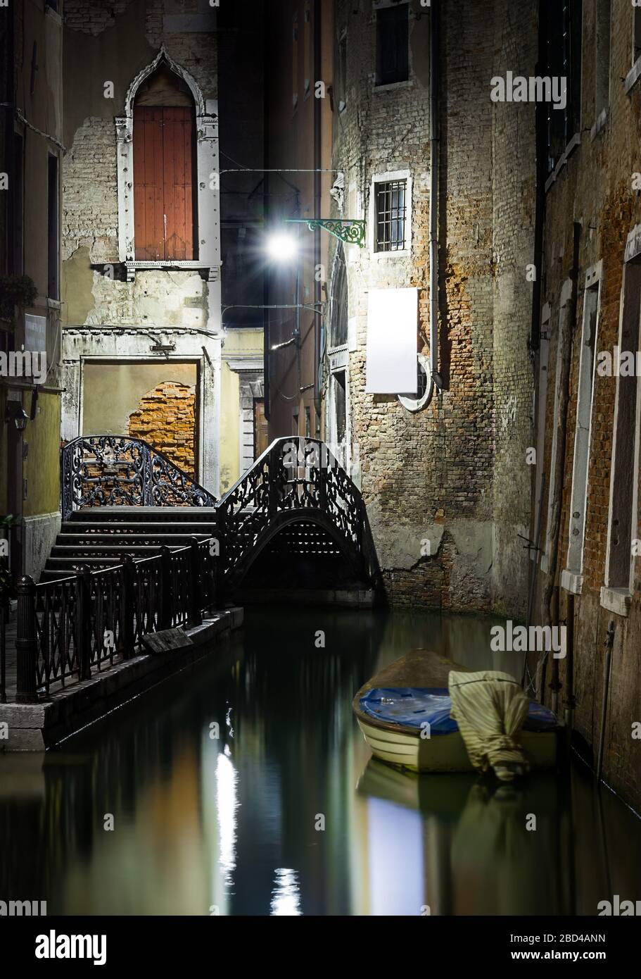 Foto nocturna de un tranquilo canal veneciano, con un barco a motor amarrado y una pasarela que conduce a un puente de hierro Foto de stock