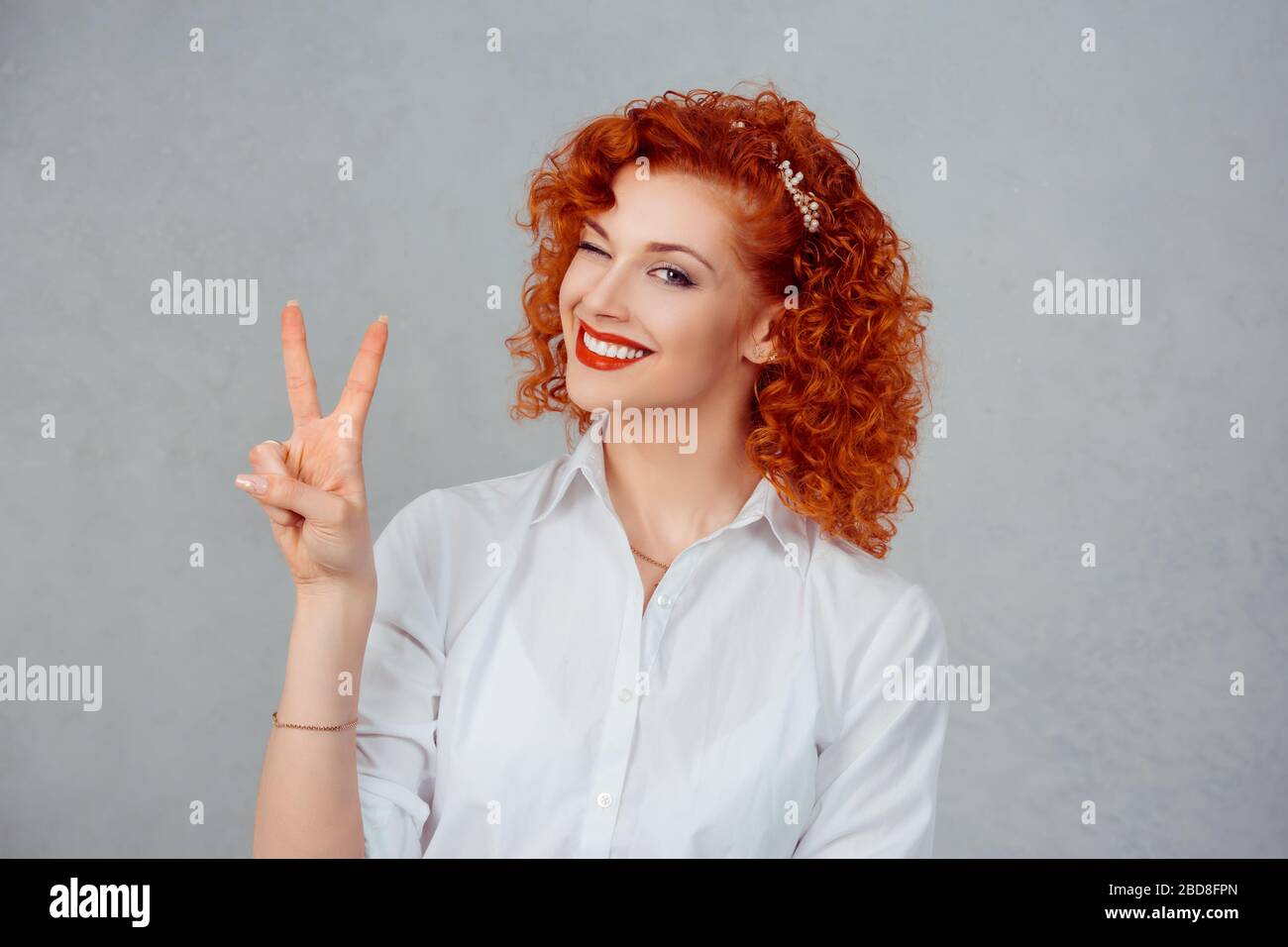 La niña atractiva está mostrando la señal de paz y sonriendo. Redhead rizado mujer de negocios guiñando y mirando la cámara de forma juguetonera. Aislado sobre fondo gris Foto de stock