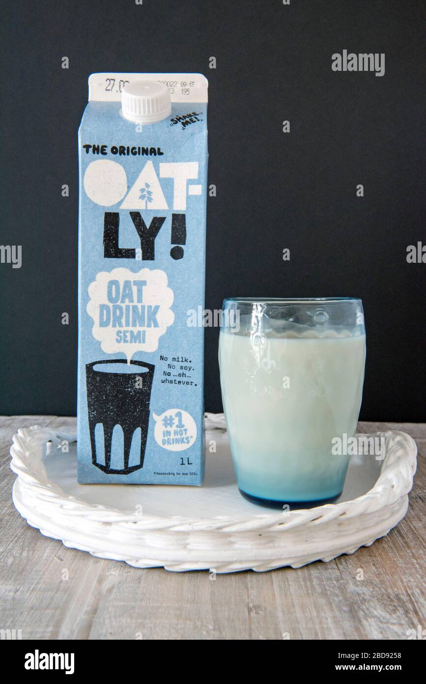 Vegan Oatly Oat leche bebida en vidrio azul vintage con el original Oatly Oat cartón de bebida detrás en bandeja blanca con fondo negro. Uso editoral onl Foto de stock