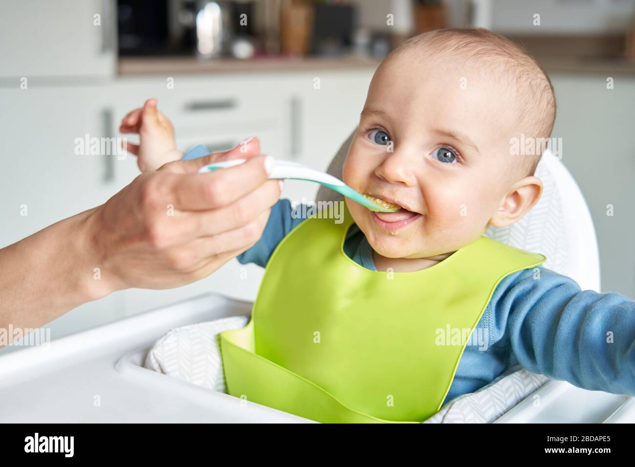 Retrato de Smiling 8 meses de niño viejo en casa en silla alta siendo alimentado de alimentos sólidos por la madre con cuchara Foto de stock