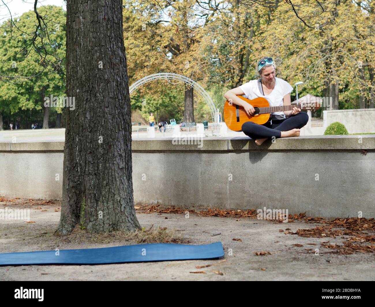 Niña sentada en una pared tocando la guitarra afuera en un día soleado en el parque con un gran tronco de árbol delante de ella. Foto de stock