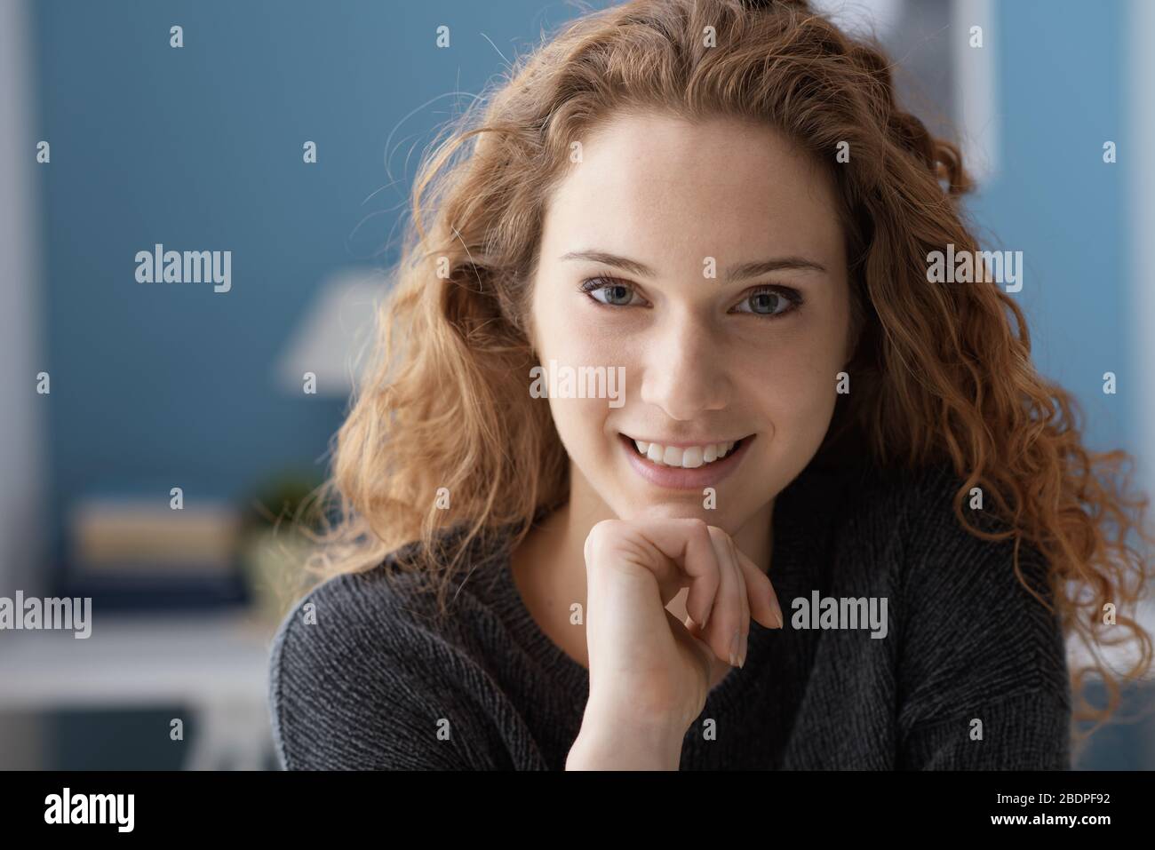 Hermosa mujer joven rizado posando en casa con la mano en la barbilla, ella está sonriendo con la cámara Foto de stock