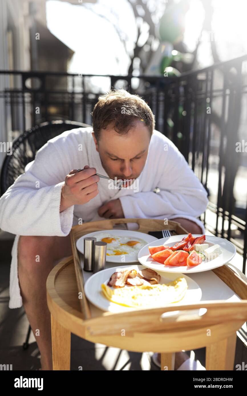 Hombre solo sentado en la silla en el balcón y comiendo sabrosos huevos con verduras por la mañana. Llevar un albornoz blanco suave. Primavera soleada y cálida Foto de stock
