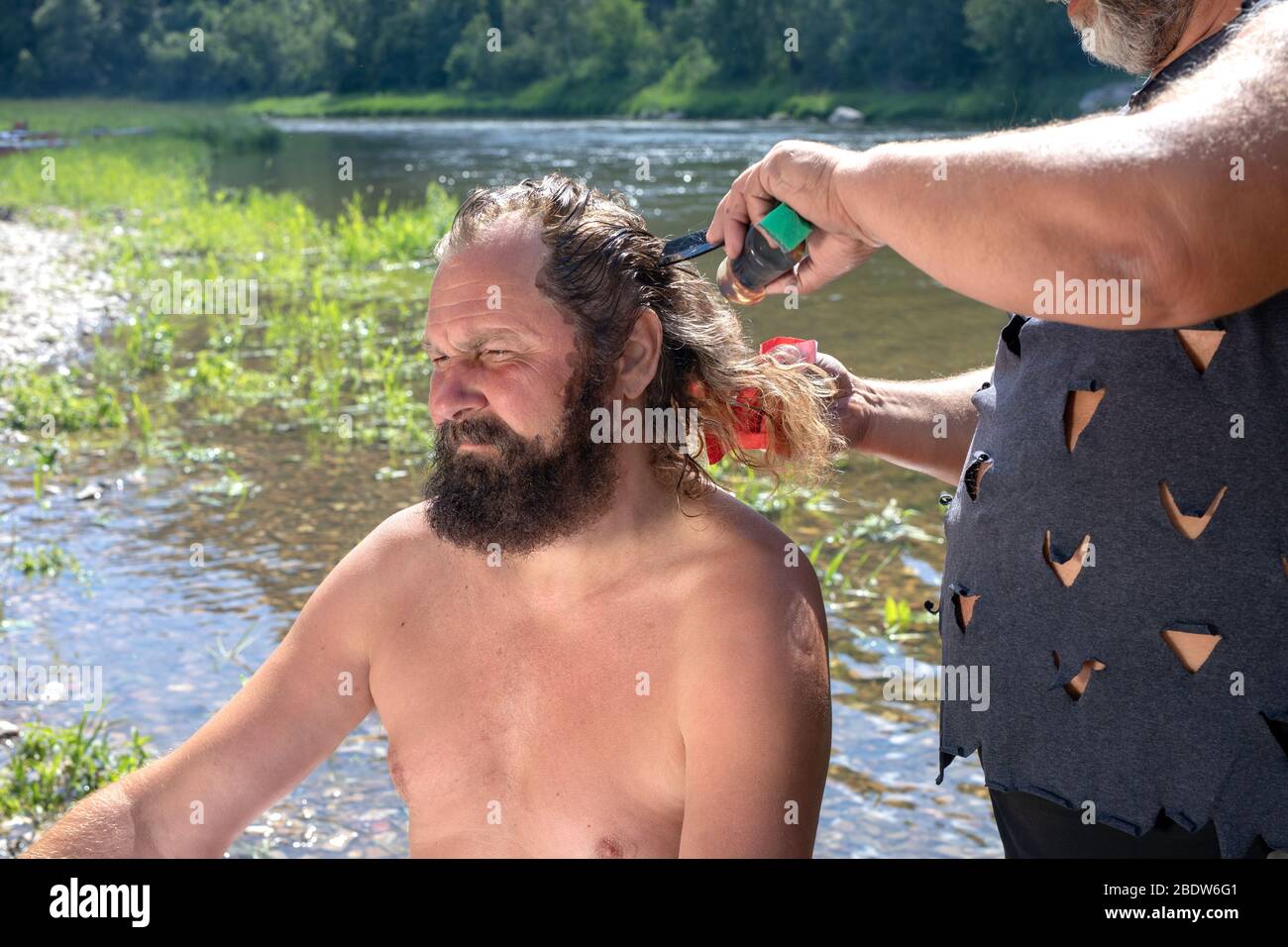 Brutales cortes de barbería o tintes pelo largo de un viejo turista sin  barba en el soleado día de verano a orillas del río bosque. Estilista,  peluquería, salón outdo Fotografía de stock -