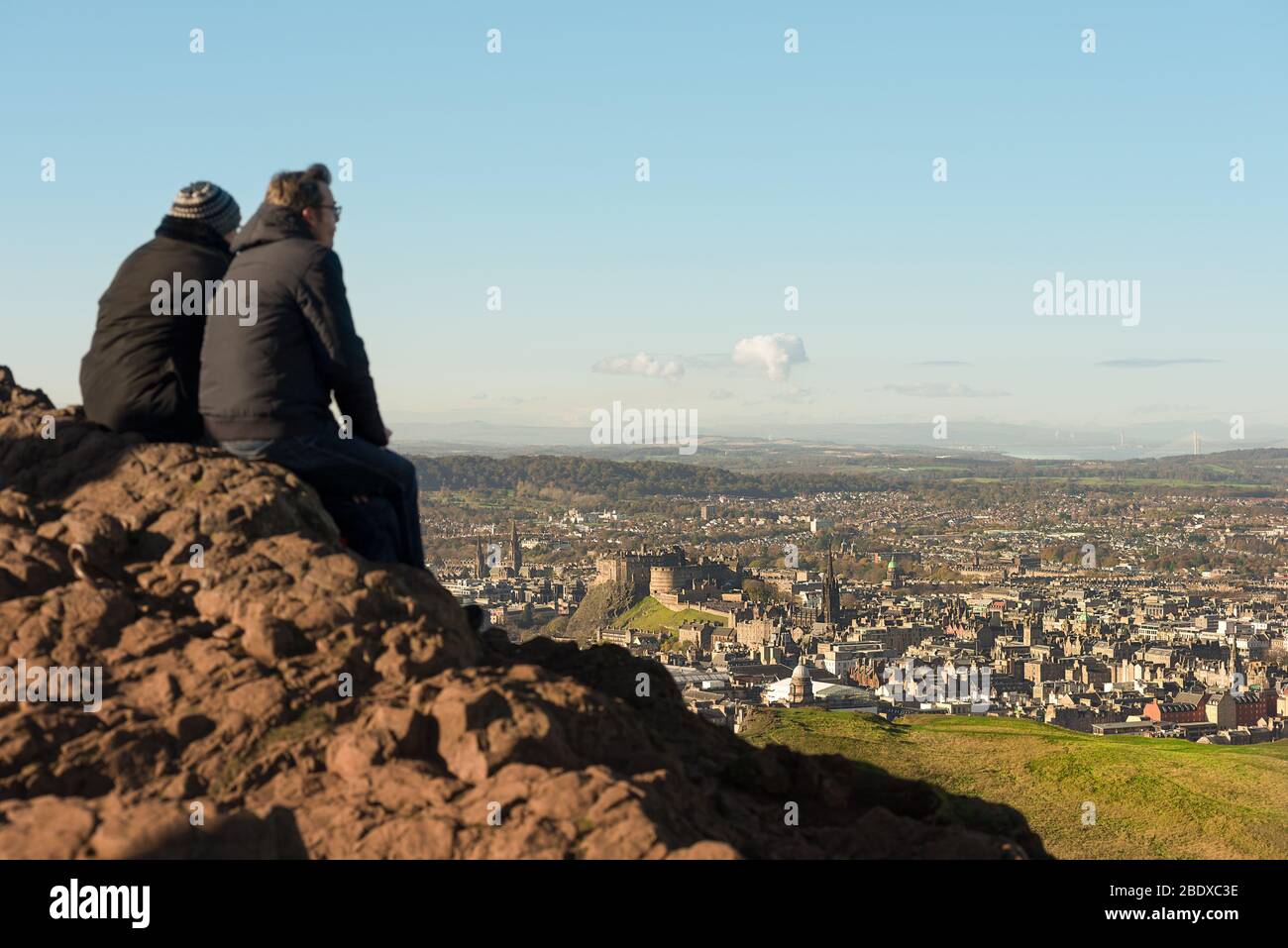 Una pareja se sienta en la parte superior de Arthurs Seat, un volcán inactivo, con vistas a la ciudad de Edimburgo y el Castillo de Edimburgo, Escocia. Foto de stock
