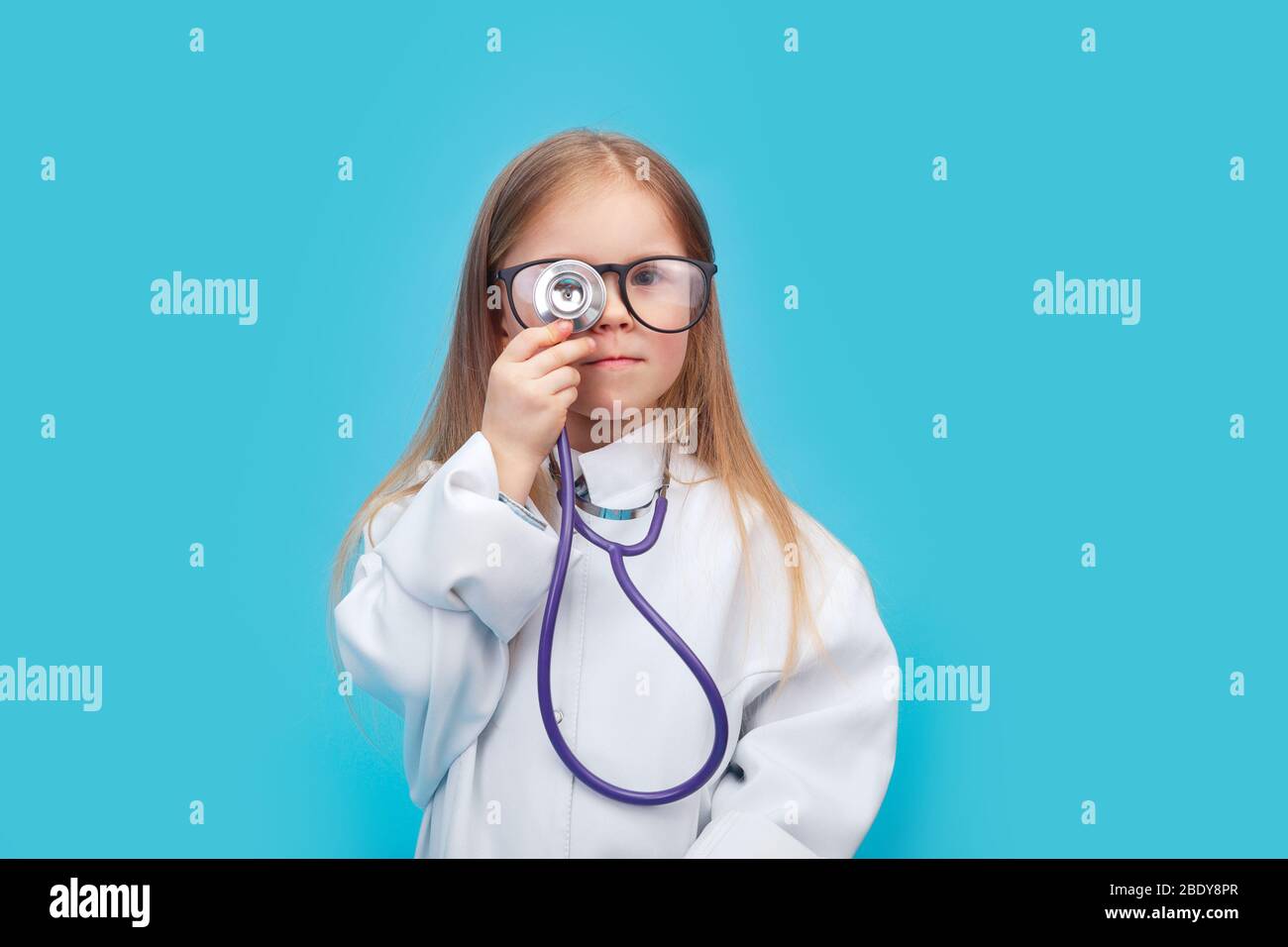 Niña sonriente con uniforme médico y gafas jugando con estetoscopio Foto de stock