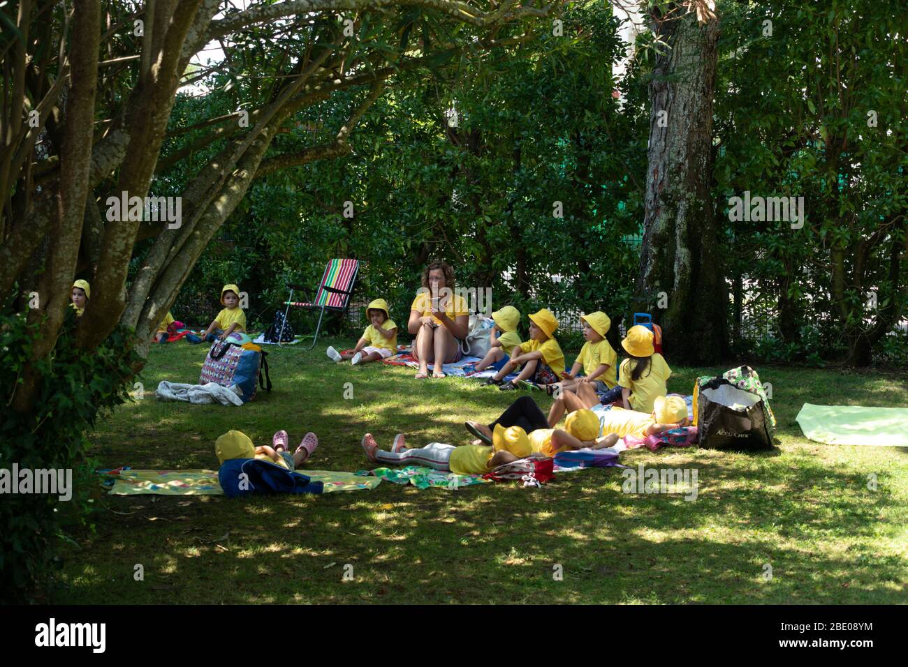 Niños sentados con sombreros amarillos y camisetas con maestro Asistir al club de verano de los niños en el hermoso jardín municipal arbolado en En Oeiras Portugal Foto de stock