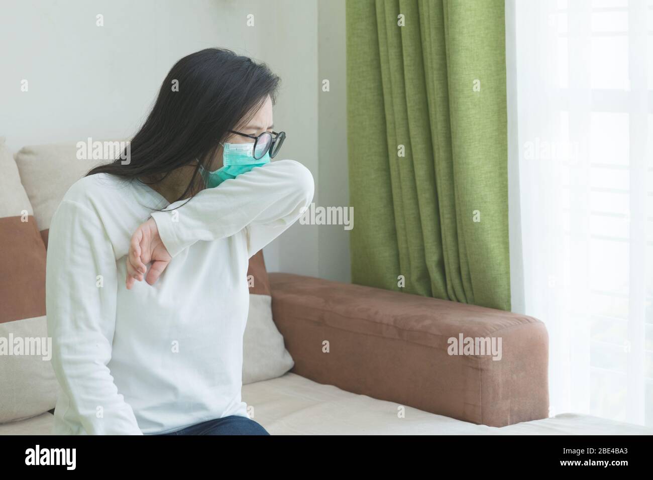 Tos de codo, estornudos de una joven hermosa mujer usando una máscara médica verde en el fondo de la sala de estar. Concepto de cuarentena de coronavirus (Covid-19). Foto de stock