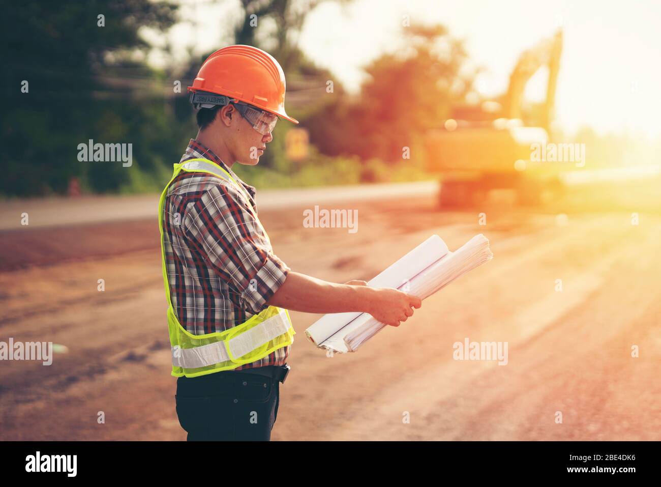 El ingeniero lleva un hellmet duro y sostiene el plano en la obra de la construcción de la carretera con la maquinaria, el concepto de seguridad primero Foto de stock