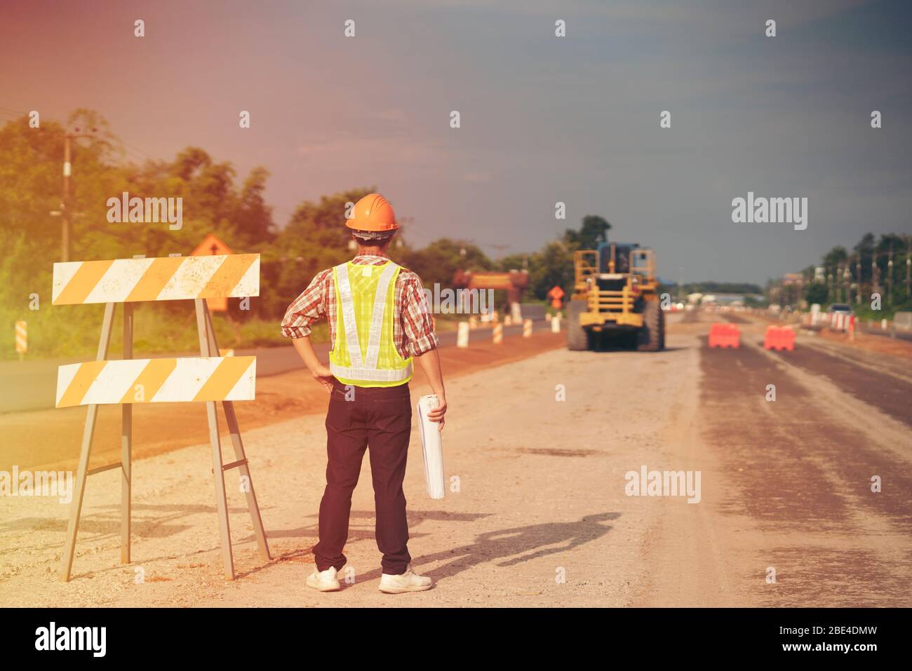 El ingeniero lleva un casco duro y un plano de sujeción en la obra de construcción de carreteras con la maquinaria, el concepto de seguridad es lo primero Foto de stock