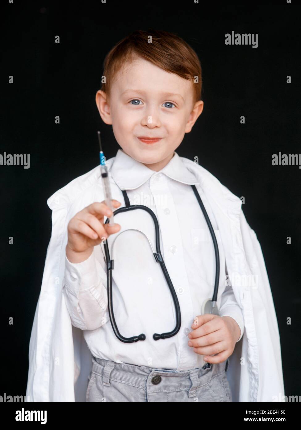 Un niño en el papel de médico. El niño elige la profesión de médico. Jeringa en sus manos sobre fondo negro Foto de stock