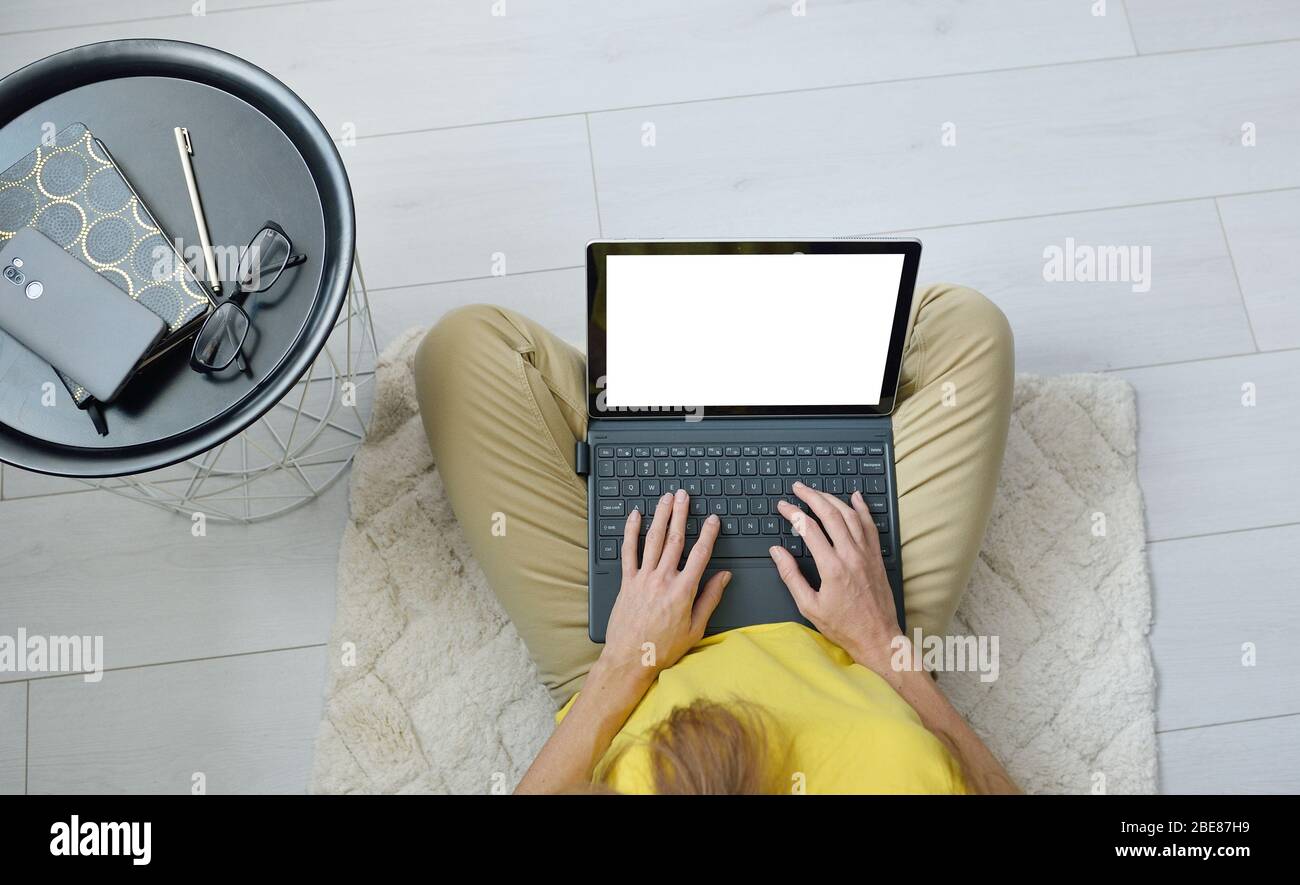 Vista superior de la mujer independiente sentada en el suelo cerca del sofá en casa y trabajando en un ordenador portátil con diseño de mascup. Mujer escribiendo en el portátil. Concepto de cuarentena Foto de stock