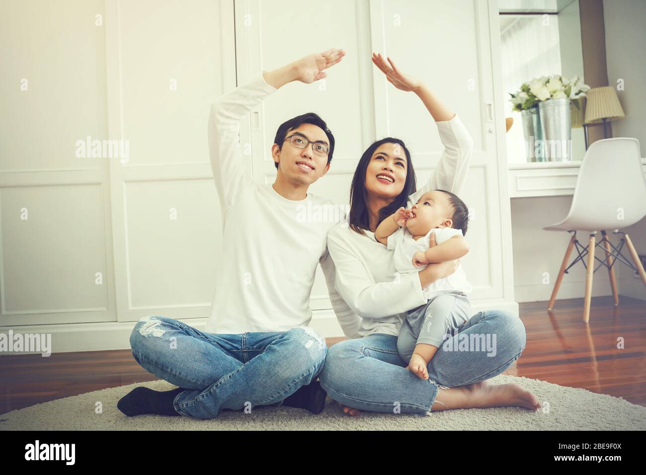 Positivo joven asiático casado pareja con un niño mirando hacia arriba y  haciendo figura de techo con las manos mientras se sienta en la alfombra en  la habitación de luz Fotografía de