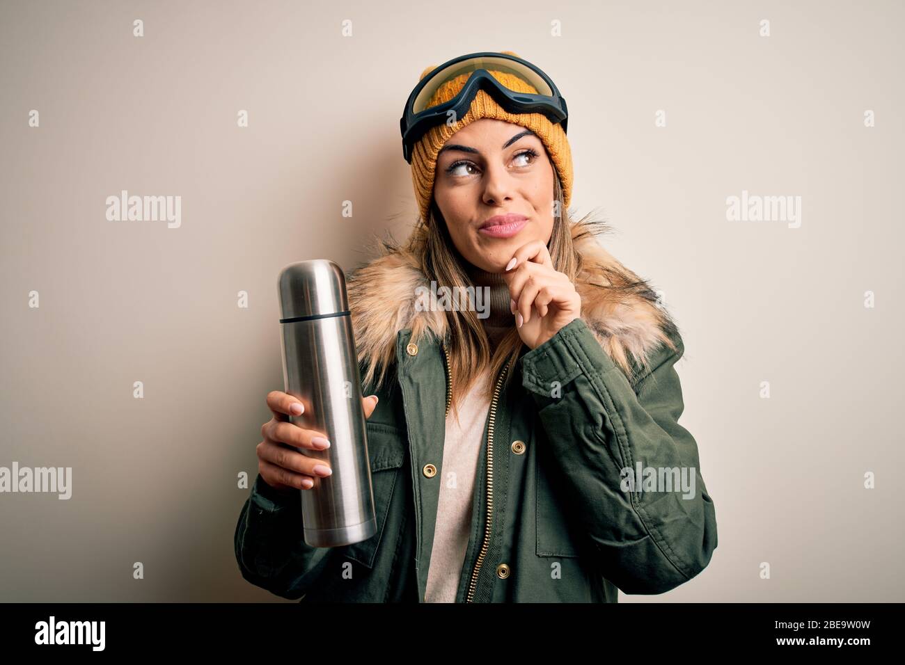 Joven hermosa morena skier mujer usando gafas de esquí tomando termo con café cara seria pensando en la pregunta, idea muy confusa Foto de stock