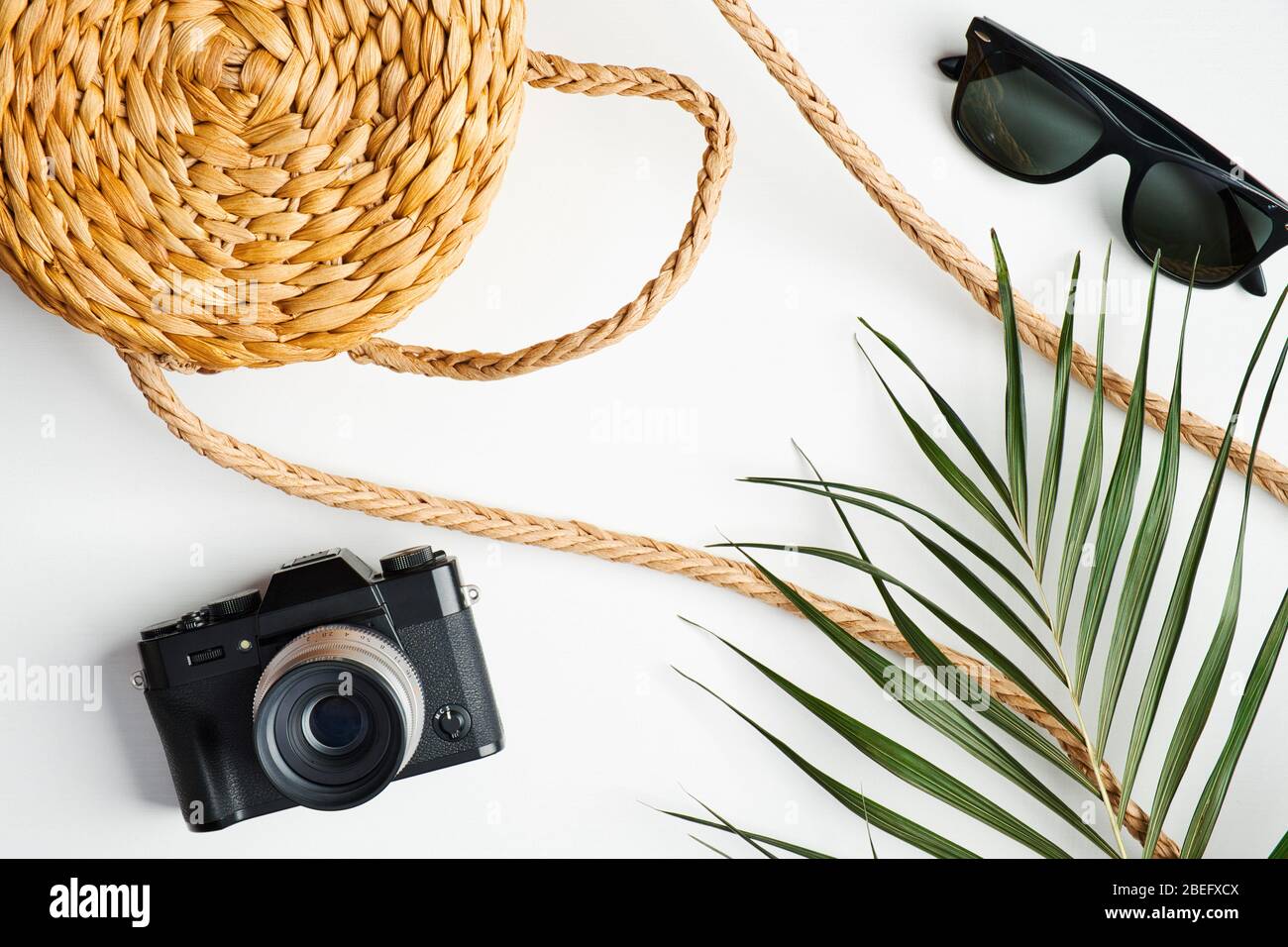 Ropa femenina de moda de verano con bolso de ratán, cámara de época, gafas de sol, hoja de palma tropical sobre mesa blanca. Concepto de viaje, vacaciones, vacaciones. FL Foto de stock