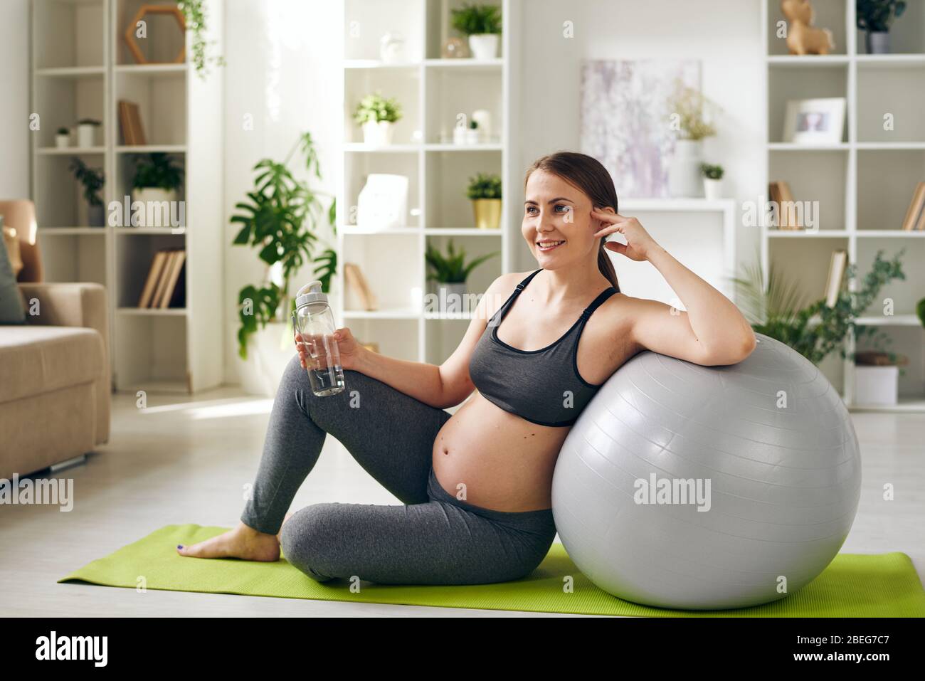 Sonriendo mujer embarazada joven y sana en ropa deportiva sentada en la esterilla de yoga y apoyada en el fitball mientras bebía agua en casa Foto de stock