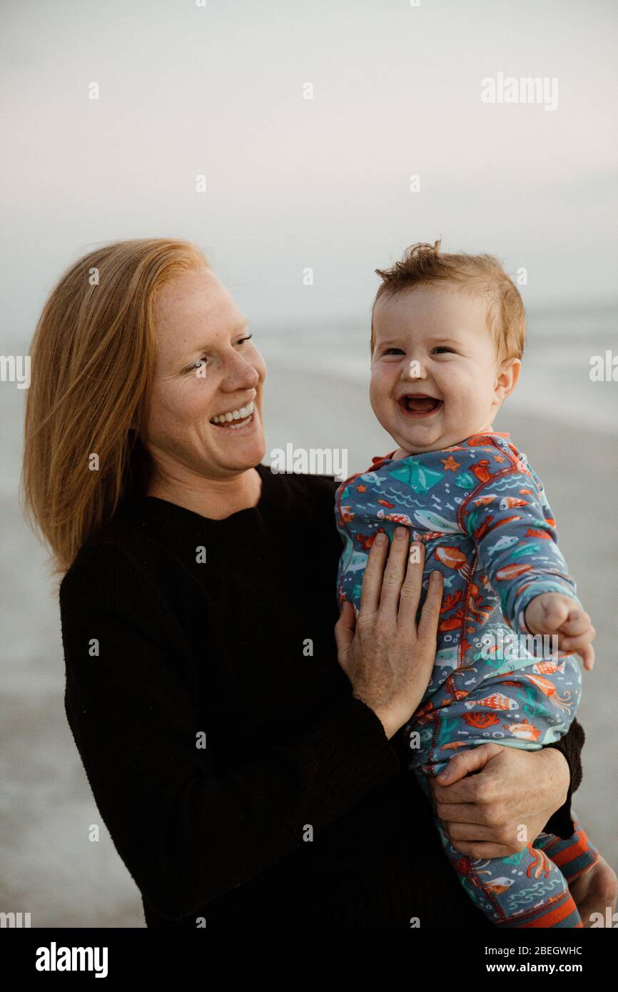 mamá soltera con pelo rojo sostiene riendo niño chubby en la playa Foto de stock