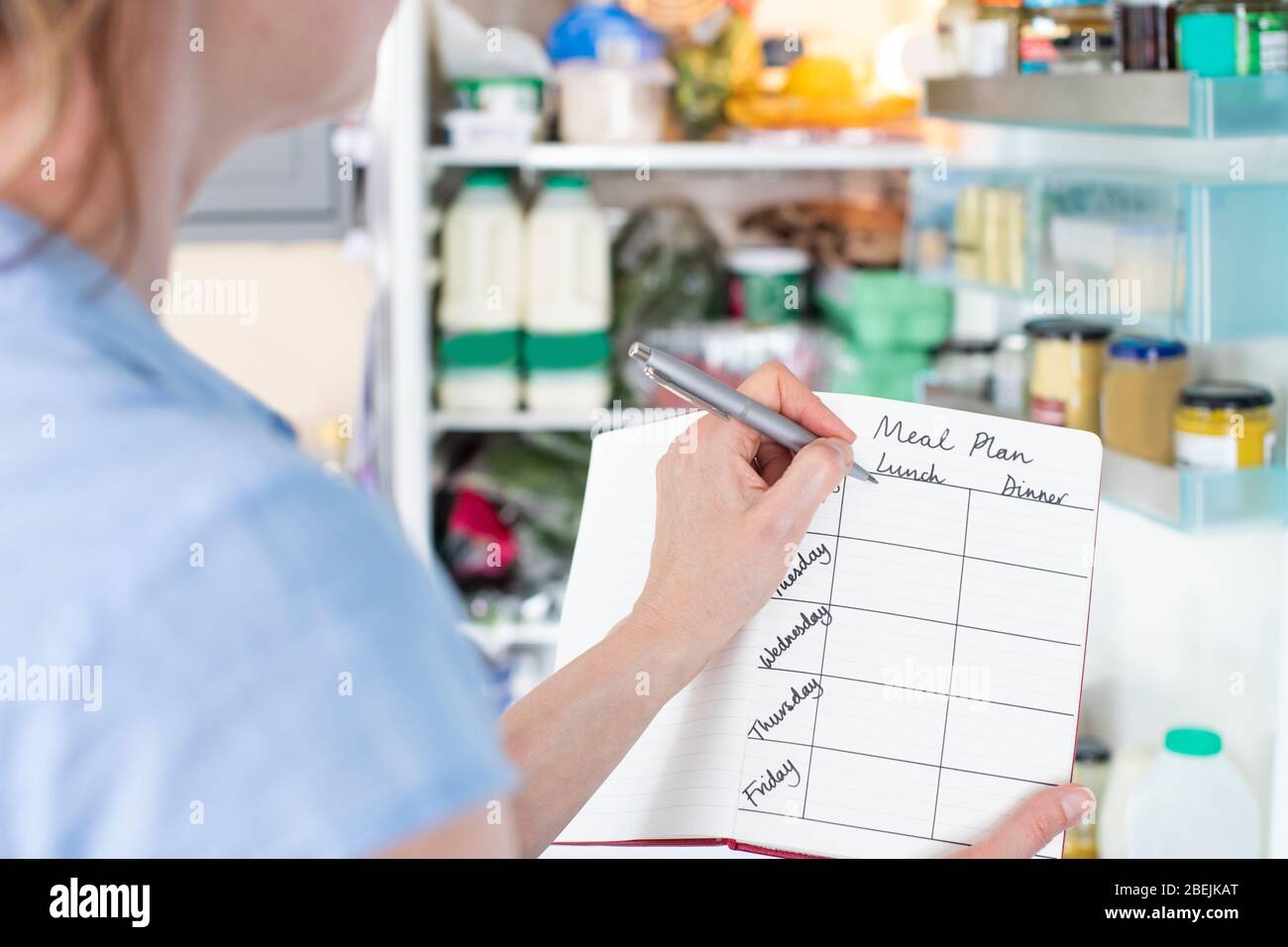 Mujer de pie frente a refrigerador en la cocina con un cuaderno de escribir el plan de comida semanal Foto de stock