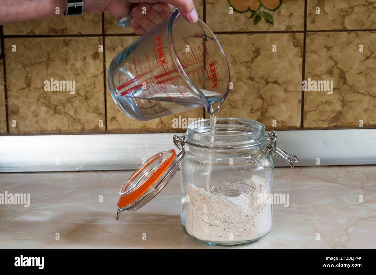 Preparación de un arrancador de masa fermentada - adición de agua a la harina en un tarro de cristal para horno. Foto de stock