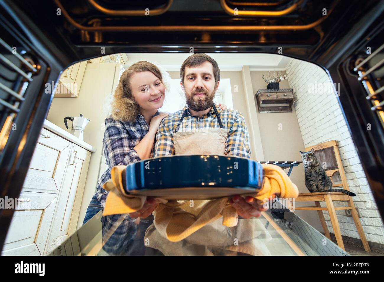 Una pareja sonriente de Europa del este cocinando en casa. Hombre sacando una tarta del horno con cocina interior y un gato en el fondo, vista desde dentro Foto de stock