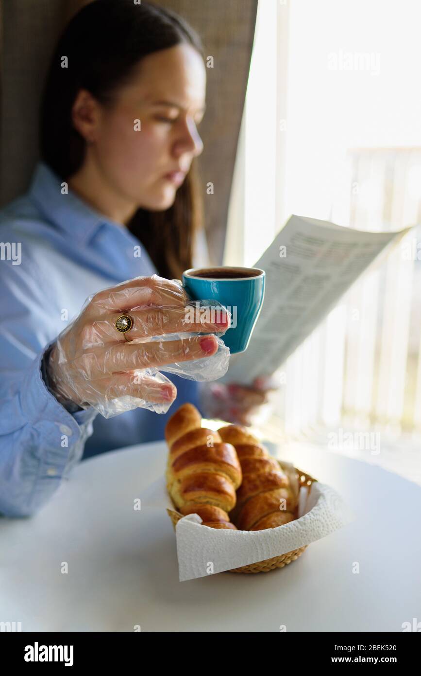 Mujer tomando el desayuno con guantes de plástico. Café y croissant, periódico. Concepto de restricciones a la cuarentena y a la pandemia Foto de stock
