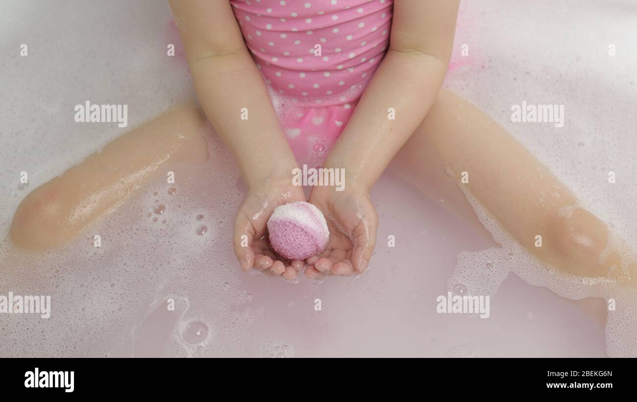 Las manos de la niña en traje de baño rosa ponen la bomba de baño en el agua  en el baño. La bola de sal del baño se disuelve en el agua